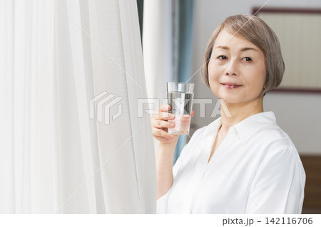 リビングの窓辺でコップで水を飲むシニア女性 リビングの窓辺でコップで水を飲むシニア女性 142116706