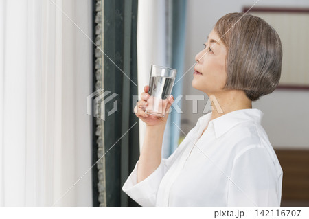 リビングの窓辺でコップで水を飲むシニア女性 142116707