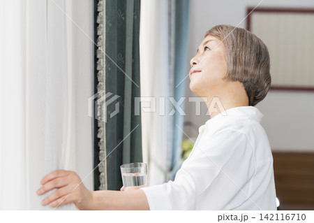 リビングの窓辺でコップで水を飲むシニア女性 リビングの窓辺でコップで水を飲むシニア女性 142116710