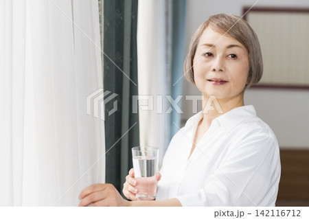 リビングの窓辺でコップで水を飲むシニア女性 142116712