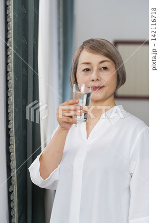 リビングの窓辺でコップで水を飲むシニア女性 142116718