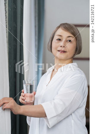 リビングの窓辺でコップで水を飲むシニア女性 リビングの窓辺でコップで水を飲むシニア女性 142116721