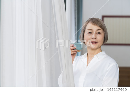 リビングの窓辺で湯呑みでお茶を飲むシニア女性 リビングの窓辺で湯呑みでお茶を飲むシニア女性 142117460