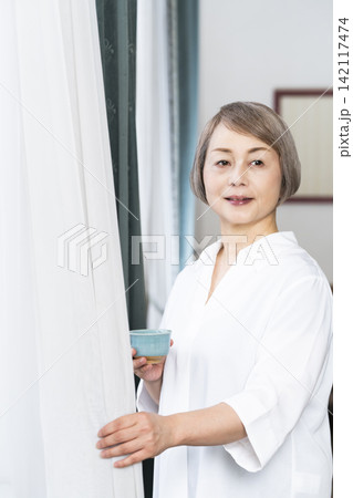 リビングの窓辺で湯呑みでお茶を飲むシニア女性 リビングの窓辺で湯呑みでお茶を飲むシニア女性 142117474