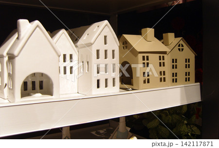 A row of decorative ceramic and matte beige miniature houses displayed on a retail shelf, designed as tea light holders with intricate cutout windows and doors. Perfect for home decor or collectibles A row of decorative ceramic and matte beige miniature houses displayed on a retail shelf, designed as tea light holders with intricate cutout windows and doors. Perfect for home decor or collectibles 142117871