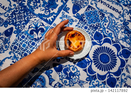 Top down food shot of tanned female hand placing one pastel de nata custard tart on white saucer on blue and white tablecloth with traditional Portuguese azulejo tile motifs with simple copy space Top down food shot of tanned female hand placing one pastel de nata custard tart on white saucer on blue and white tablecloth with traditional Portuguese azulejo tile motifs with simple copy space 142117892
