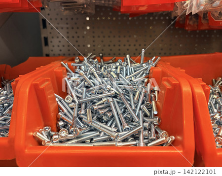 an orange bin filled with shiny silver bolts and screws in a hardware store. Plastic bags of similar hardware an orange bin filled with shiny silver bolts and screws in a hardware store. Plastic bags of similar hardware 142122101