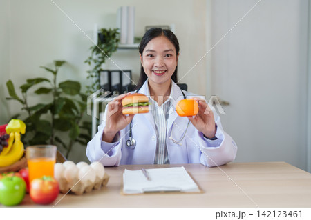 Asian nutritionist doctor holding hamburger and orange for healthy diet choice Asian nutritionist doctor holding hamburger and orange for healthy diet choice 142123461