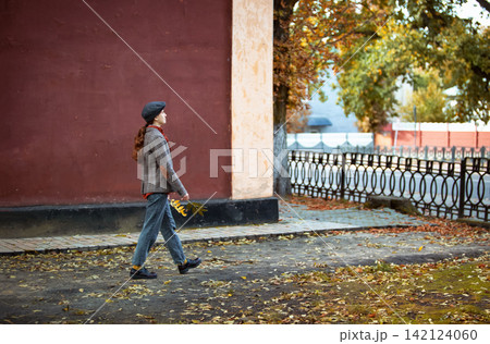 Young stylish woman wearing a flat cap and tweed jacket holding autumn leaves while walking along a city street next to a red building wall. Autumn street fashion, red brick wall, fall foliage leaves Young stylish woman wearing a flat cap and tweed jacket holding autumn leaves while walking along a city street next to a red building wall. Autumn street fashion, red brick wall, fall foliage leaves 142124060