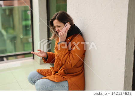 Woman in orange coat sitting beside wall and talking on smartphone while gesturing with open hand indoors near bright window, showing concern and waiting for response Woman in orange coat sitting beside wall and talking on smartphone while gesturing with open hand indoors near bright window, showing concern and waiting for response 142124622