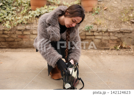 Young woman crouching outdoors while checking transparent backpack beside stone wall, wearing furry coat, long braid, gloves, and boots in soft natural light with autumn tones Young woman crouching outdoors while checking transparent backpack beside stone wall, wearing furry coat, long braid, gloves, and boots in soft natural light with autumn tones 142124623