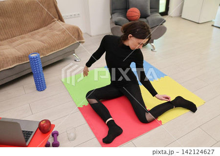 Woman stretching on colorful exercise mat in bright home living room, surrounded by fitness props, foam roller, dumbbell, laptop, apple, glass, and basketball for active wellness routine Woman stretching on colorful exercise mat in bright home living room, surrounded by fitness props, foam roller, dumbbell, laptop, apple, glass, and basketball for active wellness routine 142124627
