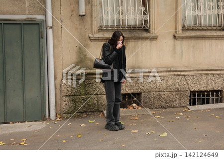 Woman in black coat standing beside weathered city wall, holding tissue to face near quiet sidewalk with autumn leaves, worn windows, old doorway, and muted urban textures 142124649