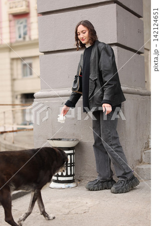 Young woman in dark casual clothing stands beside gray stone wall on city sidewalk, looking down while holding tissue near stray dog passing through foreground, with woman, dog, urban, sidewalk detail Young woman in dark casual clothing stands beside gray stone wall on city sidewalk, looking down while holding tissue near stray dog passing through foreground, with woman, dog, urban, sidewalk detail 142124651