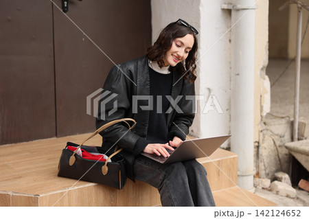 Young woman sitting on outdoor steps using laptop beside handbag, smiling while working in casual black outfit near weathered urban wall, relaxed modern lifestyle and remote work concept Young woman sitting on outdoor steps using laptop beside handbag, smiling while working in casual black outfit near weathered urban wall, relaxed modern lifestyle and remote work concept 142124652