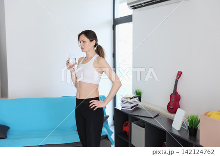 Young woman standing in bright modern living room holding glass of water beside blue sofa, shelves with books, small plants, ukulele, laptop and window light in calm home interior Young woman standing in bright modern living room holding glass of water beside blue sofa, shelves with books, small plants, ukulele, laptop and window light in calm home interior 142124762