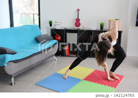 Woman performing deep side stretch on colorful foam mat in bright modern living room with sofa, shelving, indoor plants, and casual home exercise setup, with yoga, stretching, fitness, exercise detail Woman performing deep side stretch on colorful foam mat in bright modern living room with sofa, shelving, indoor plants, and casual home exercise setup, with yoga, stretching, fitness, exercise detail 142124765