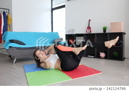 Woman doing bicycle crunch exercise on colorful mat in bright home living room with sofa, shelves, clothes rack, fitness accessories, and natural daylight creating healthy active lifestyle scene Woman doing bicycle crunch exercise on colorful mat in bright home living room with sofa, shelves, clothes rack, fitness accessories, and natural daylight creating healthy active lifestyle scene 142124766