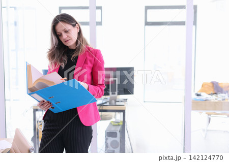 Businesswoman in pink blazer reviewing documents in bright modern office while holding blue folder and standing near computer workstation, focused on paperwork and office tasks 142124770