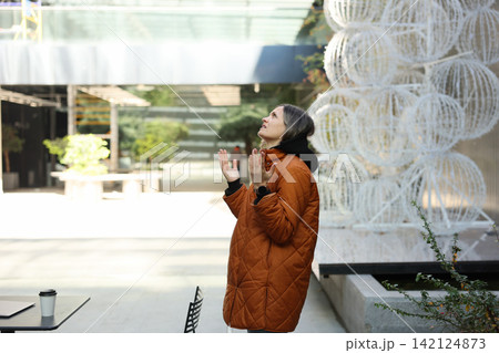Thoughtful woman in orange quilted coat looking upward with raised hands beside white circular wire sculpture in bright modern courtyard with soft daylight, urban calm, reflective mood and clean Thoughtful woman in orange quilted coat looking upward with raised hands beside white circular wire sculpture in bright modern courtyard with soft daylight, urban calm, reflective mood and clean 142124873