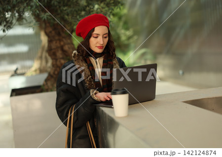 Young woman in red beret using laptop outdoors beside coffee cup, wearing winter coat and scarf, focused on work at modern urban terrace with soft natural light and blurred greenery Young woman in red beret using laptop outdoors beside coffee cup, wearing winter coat and scarf, focused on work at modern urban terrace with soft natural light and blurred greenery 142124874