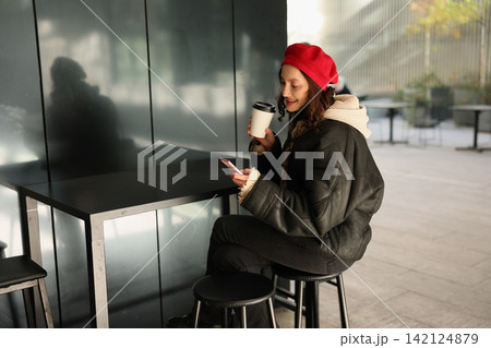 Young woman in red beret sits at modern cafe table, smiling at smartphone while holding takeaway coffee cup, wearing dark coat and scarf beside reflective wall in soft daylight Young woman in red beret sits at modern cafe table, smiling at smartphone while holding takeaway coffee cup, wearing dark coat and scarf beside reflective wall in soft daylight 142124879