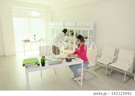 Bright minimal office with young woman working at desk while colleague checks cabinet supplies, white modern workspace with chairs, binders, desk items, soft daylight, calm clean interior Bright minimal office with young woman working at desk while colleague checks cabinet supplies, white modern workspace with chairs, binders, desk items, soft daylight, calm clean interior 142125042