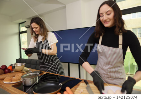 Two women preparing breakfast together in bright modern kitchen, wearing aprons and gloves while cooking with fresh ingredients on wooden counter near stove and window light Two women preparing breakfast together in bright modern kitchen, wearing aprons and gloves while cooking with fresh ingredients on wooden counter near stove and window light 142125074