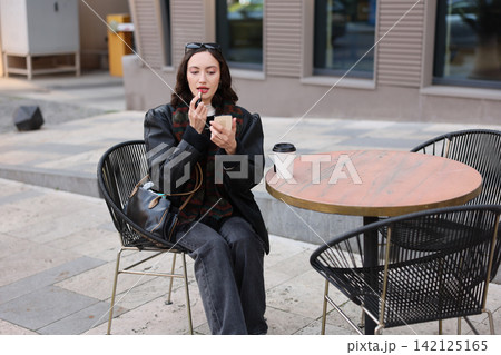 Young woman applying lipstick while seated outdoors at urban cafe table with handbag and coffee cup beside her in soft daylight, stylish casual fashion portrait Young woman applying lipstick while seated outdoors at urban cafe table with handbag and coffee cup beside her in soft daylight, stylish casual fashion portrait 142125165