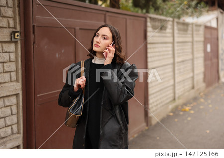 Young woman in black coat holding smartphone near residential wall, standing outdoors on quiet street with thoughtful expression, shoulder bag, soft natural light and shallow focus Young woman in black coat holding smartphone near residential wall, standing outdoors on quiet street with thoughtful expression, shoulder bag, soft natural light and shallow focus 142125166