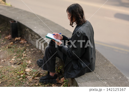 Young woman sitting on stone wall outdoors writing notes in small notebook beside quiet road, wearing black jacket and shoes, focused on journaling in calm autumn setting Young woman sitting on stone wall outdoors writing notes in small notebook beside quiet road, wearing black jacket and shoes, focused on journaling in calm autumn setting 142125167