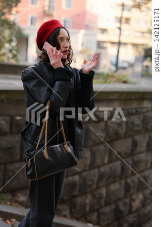 Young woman in red beret talking on smartphone outdoors, showing surprised expression and gesturing with hand while standing near blurred city building and stone wall Young woman in red beret talking on smartphone outdoors, showing surprised expression and gesturing with hand while standing near blurred city building and stone wall 142125171