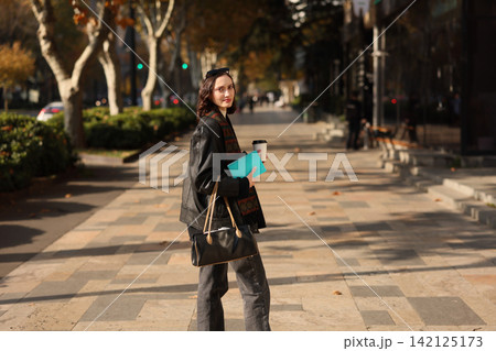 Young woman on sunlit city sidewalk holding coffee cup, notebook, and handbag, looking at camera with calm expression amid autumn trees and soft urban background blur 142125173