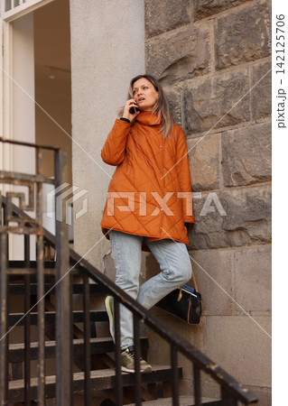 Woman in orange quilted coat stands on stone stairway, talking on smartphone beside textured wall, wearing jeans and sneakers, with calm urban portrait styling and vertical composition 142125706
