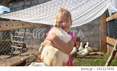 Young girl smiling and embracing a large white duck outdoors in a rustic farm pen, experiencing childhood joy, connection with animals, and rural life Young girl smiling and embracing a large white duck outdoors in a rustic farm pen, experiencing childhood joy, connection with animals, and rural life 142127838