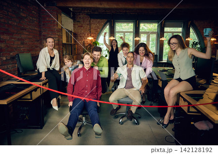 Office colleagues race chairs toward red finish marker while team cheers behind them. Office colleagues race chairs toward red finish marker while team cheers behind them. 142128192