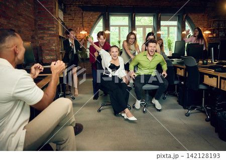 Office team cheering for colleagues competing in chair race during company party Office team cheering for colleagues competing in chair race during company party 142128193