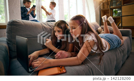 Two school-age girls browsing laptop content together while lying on sofa 142128200