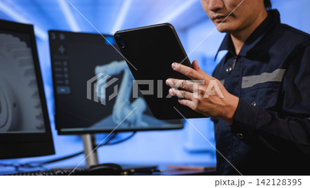 Technician using a tablet while working with robot arm screens in a blue-lit industrial control room Technician using a tablet while working with robot arm screens in a blue-lit industrial control room 142128395