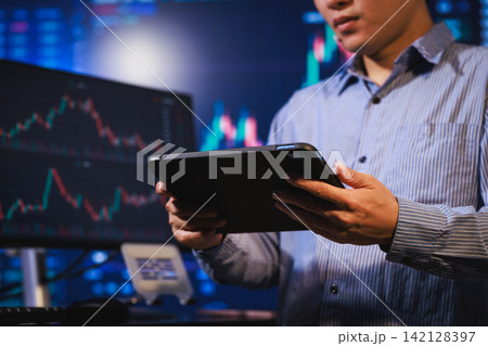 Man holding a tablet in front of computer screens with stock charts in a modern trading office. Man holding a tablet in front of computer screens with stock charts in a modern trading office. 142128397