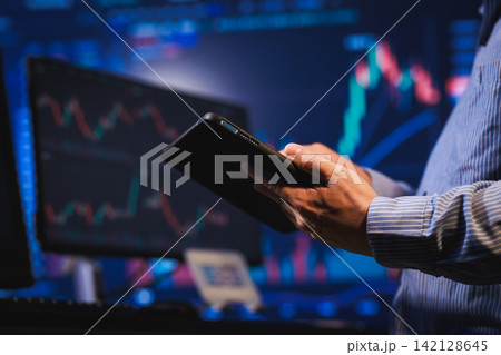 Person holding a tablet in front of stock market charts and trading screens, suggesting finance, investment, analysis, and digital business work. Person holding a tablet in front of stock market charts and trading screens, suggesting finance, investment, analysis, and digital business work. 142128645