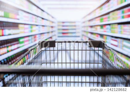 Shopping cart in a grocery store aisle with blurred shelves and bright supermarket lights, a wide selection of products in a clean retail setting Shopping cart in a grocery store aisle with blurred shelves and bright supermarket lights, a wide selection of products in a clean retail setting 142128682