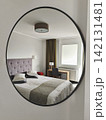 Oval wall mirror reflecting modern bedroom with double bed, upholstered headboard, window, desk and soft neutral decor 142131481