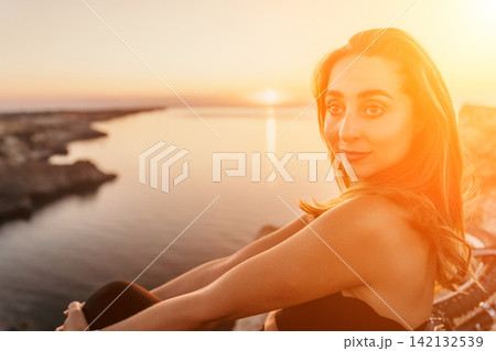 Woman sunset ocean Beautiful young woman sitting on a cliff overlooking the calm ocean during a vibrant golden hour sunset 142132539