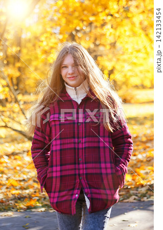Girl walks through a park with autumn leaves on the ground during a sunny day in a bright plaid jacket 142133454