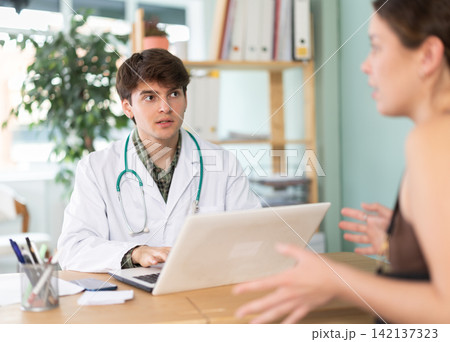 Experienced young male doctor wearing uniform with stethoscope doing check up of sick and health young girl patient sitting with laptop at table in medical office at hospital Experienced young male doctor wearing uniform with stethoscope doing check up of sick and health young girl patient sitting with laptop at table in medical office at hospital 142137323