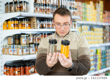 Man with glasses carefully selects jars of spices in the grocery department of hypermarket Man with glasses carefully selects jars of spices in the grocery department of hypermarket 142137494