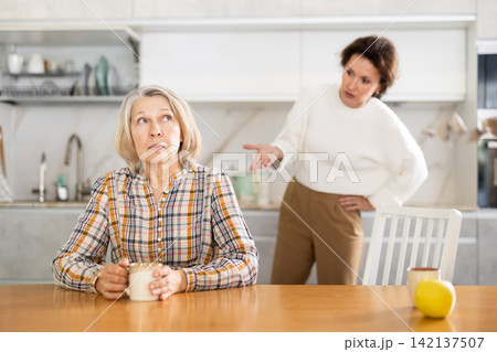 Upset old woman sitting at the kitchen table with her back to woman quarreling to her Upset old woman sitting at the kitchen table with her back to woman quarreling to her 142137507