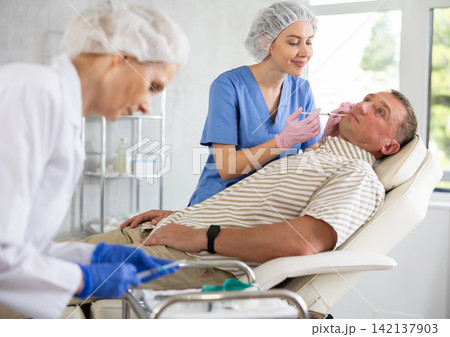 Young woman doctor gives injection of botulinum toxin in lip area to man, mature female nurse assist Young woman doctor gives injection of botulinum toxin in lip area to man, mature female nurse assist 142137903