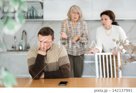 Old and middle-aged woman quarreling angrily to man sitting at the kitchen table Old and middle-aged woman quarreling angrily to man sitting at the kitchen table 142137991